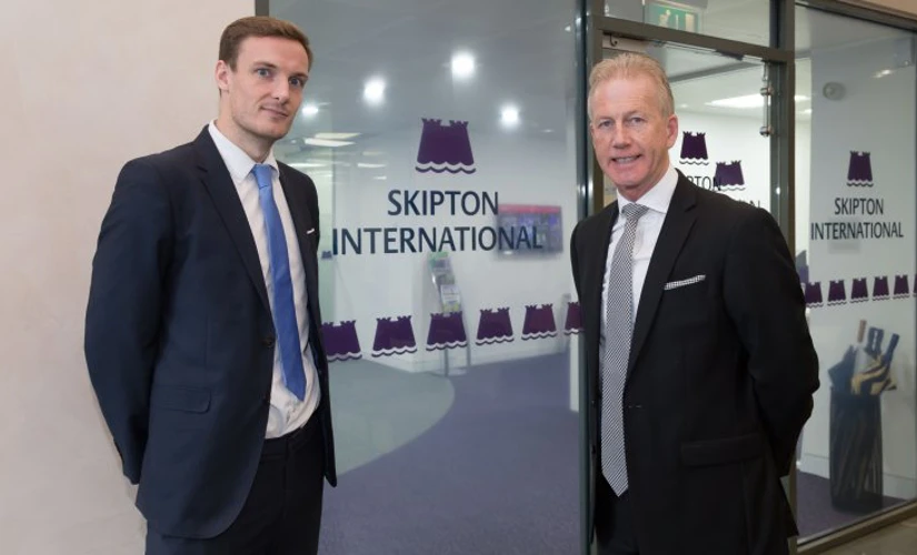 Two men smiling Infront of Skipton logo